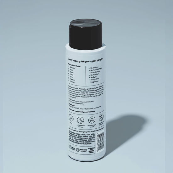 A bottle of Odele Curl Defining No-Lather Shampoo stands upright against a plain, light blue backdrop with the back of the label facing the camera.