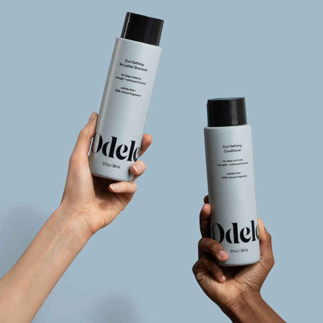 Two hands hold a bottle of Odele Beauty Curl Defining No-Lather Shampoo and Curl Defining Conditioner in front of a plain, light blue background.