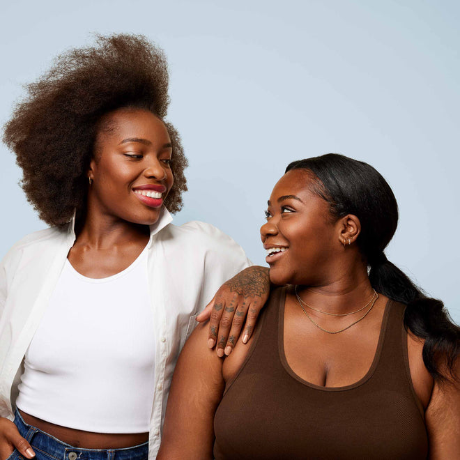 Two medium brown-skinned women pose while smiling at eachother against a plain, light blue background.