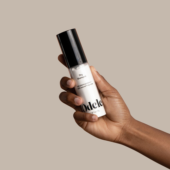 A hand holds a bottle of Odele Dry Shampoo against a beige background.