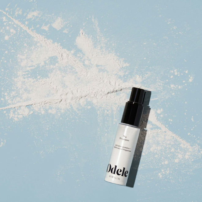 A bottle of Odele Dry Shampoo sits on a light blue surface, surrounded by an explosion of powder.