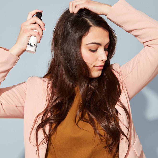 A woman with long brown hair in a pink blazer sprays Odele Dry Shampoo onto her hair in front of a plain, light blue background.