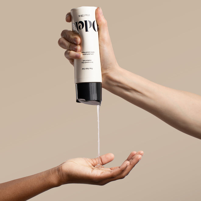 A hand squeezes a bottle of Odele Flex Hair Gel onto another’s palm in front of a plain beige background.
