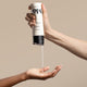 A hand squeezes a bottle of Odele Flex Hair Gel onto another’s palm in front of a plain beige background.