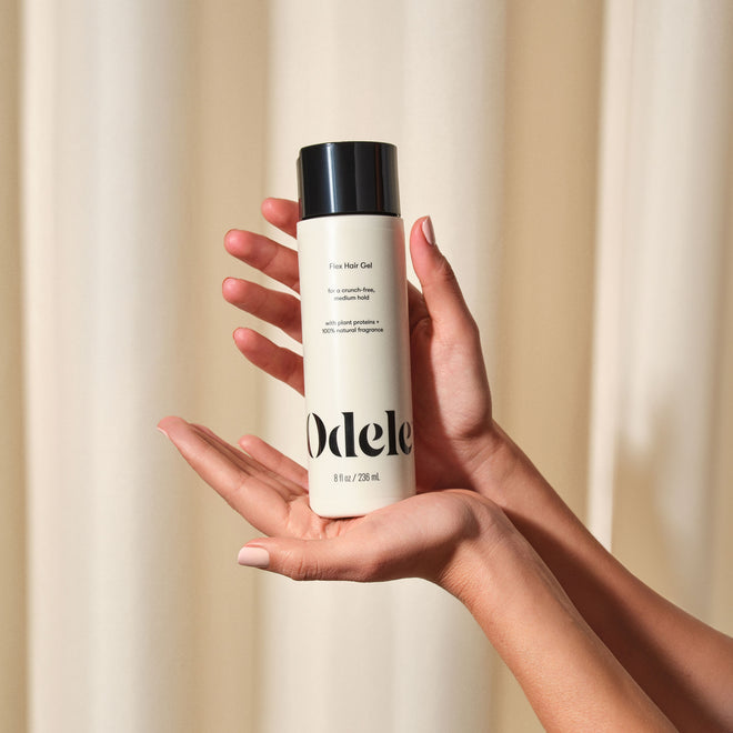 Two hands hold a bottle of Odele Flex Hair Gel in front of a soft beige curtain.