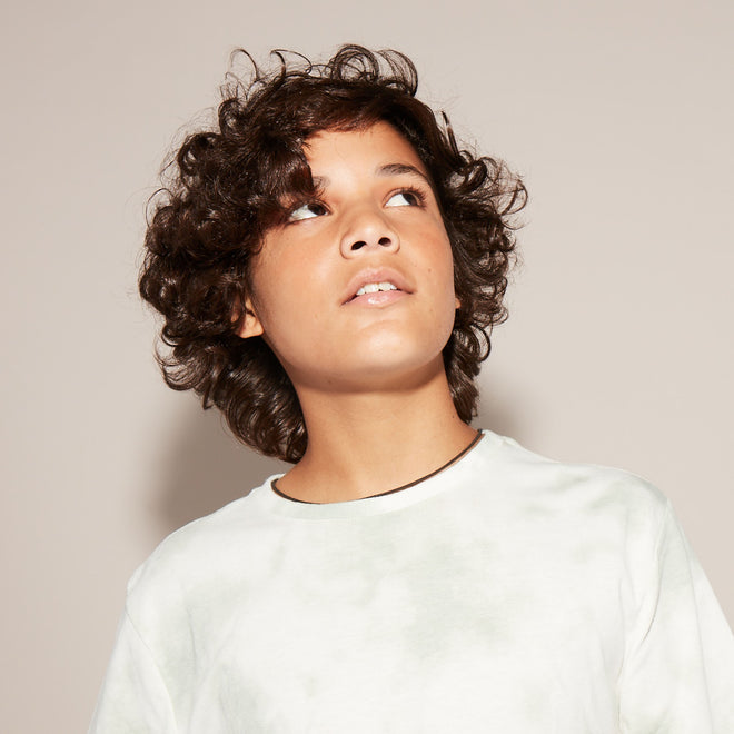 A young boy with light brown skin and curly brown hair gazes upward in front of a plain beige background.