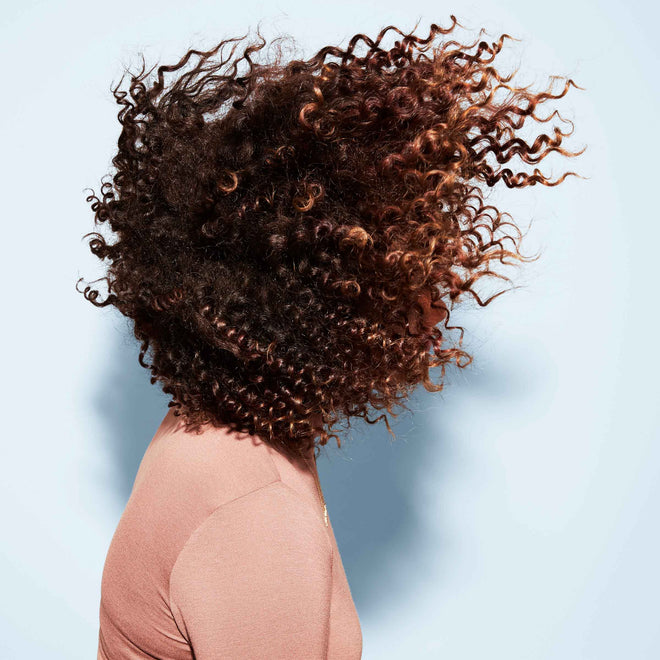 A person with voluminous curls in a light pink top looks away from the camera as their hair fans out around their head.