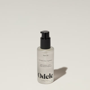 A bottle of Odele Hair Oil stands upright against a plain, light-colored backdrop with the front of the label facing the camera.