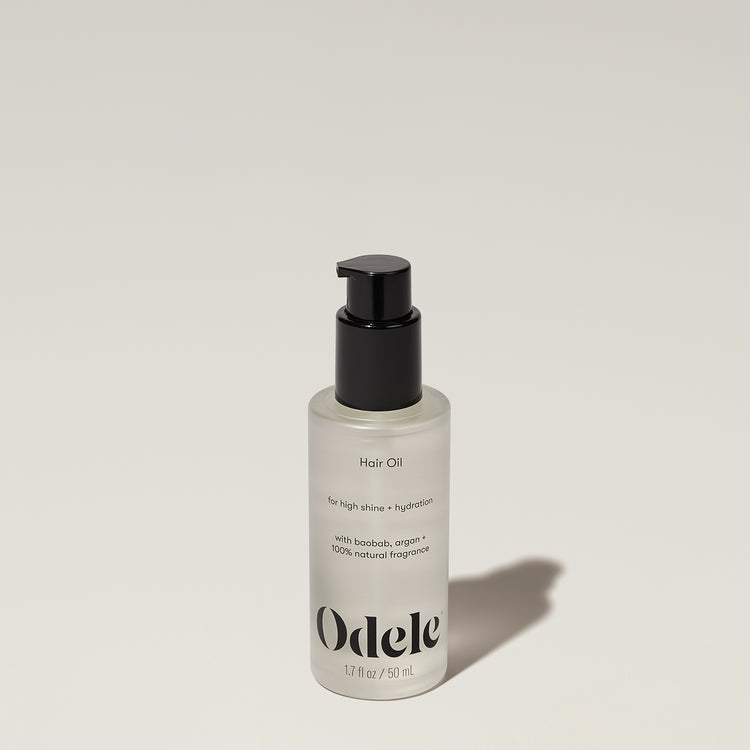 A frosted 1.7 fl oz (50 mL) bottle of Odele Beauty Hair Oil Mini with a black pump sits on a light surface, shadowed. The label notes high shine and hydration powered by nourishing argan oils.