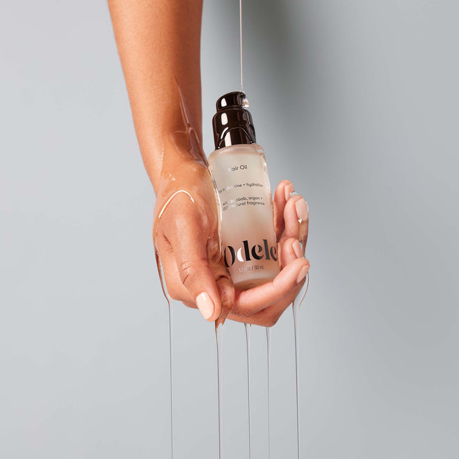 A hand holds Odele Beautys Hair Oil Mini, with the lightweight oil streaming down the wrist and fingers, creating a high-shine effect against a light gray background.