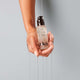 A hand dripping with oil holds a bottle of Odele Hair Oil in front of a plain, light-colored background.