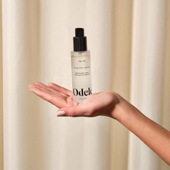 A pale-skinned hand holds a bottle of Odele Hair Oil in front of a neutral beige curtain backdrop.