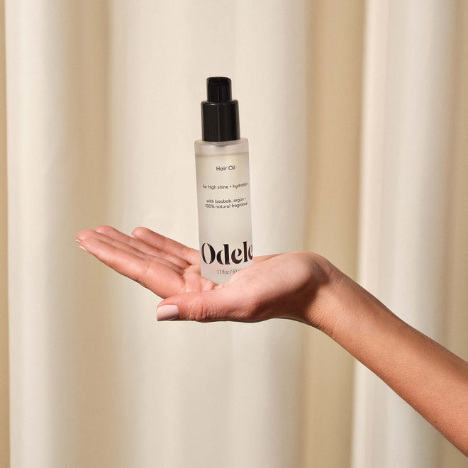 A hand with light skin tone holds a clear bottle of Odele Beauty Hair Oil Mini with a black pump, in front of a neutral beige curtain backdrop.