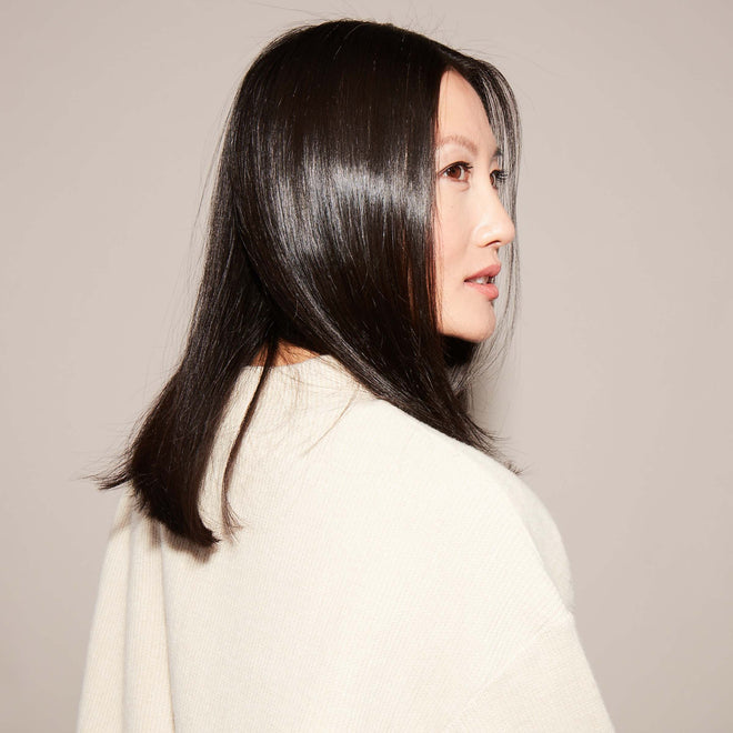 A woman with straight, high-shine black hair in a light beige top looks over her shoulder against a beige background, her style enhanced by Odele Beautys Hair Oil Mini.