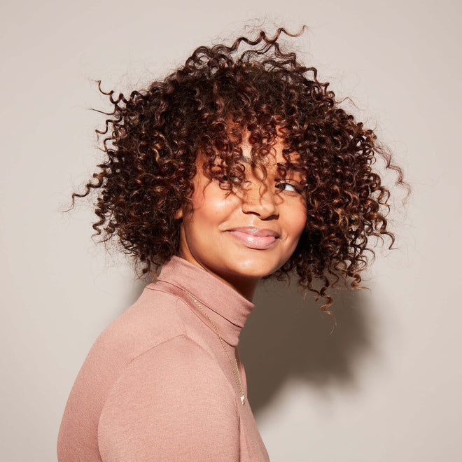 A smiling woman with short, curly hair styled using the Odele Beauty Long Live Bundle wears a light pink turtleneck, standing against a plain light background and looking slightly to the side.