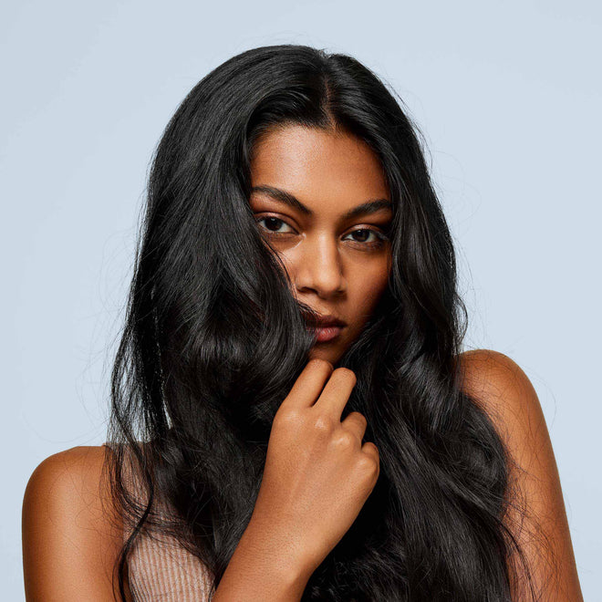 A woman with long, wavy black hair partially covering her face gazes confidently at the camera against a light blue background—her textured waves styled to perfection with Odele Beauty’s Long Live Bundle. She holds a strand near her chin.