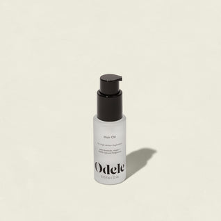 A bottle of Odele Hair Oil Mini with a black pump rests on a light off-white background, casting a soft shadow. The label highlights shine, hydration, and frizz control from nourishing argan oil and notes its natural fragrance.