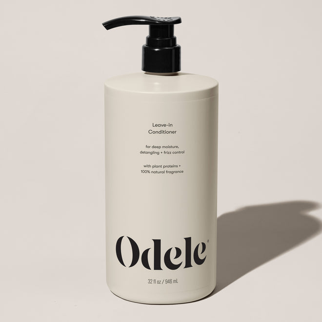 A beige bottle of Odele Leave-in Conditioner Bulk with a black pump top rests on a light surface. The label features detangling, plant proteins, 100% natural fragrance, and a 32 oz (946 ml) size.