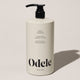 A beige bottle of Odele Leave-in Conditioner Bulk with a black pump top rests on a light surface. The label features detangling, plant proteins, 100% natural fragrance, and a 32 oz (946 ml) size.