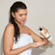 A young woman with wet hair, wrapped in a white towel, dispenses Odele Leave-in Conditioner into her hand against a plain light background.