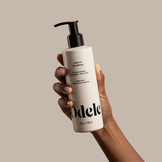 A brown skinned hand holds a bottle of Odele Leave-in Conditioner in front of a light beige background.