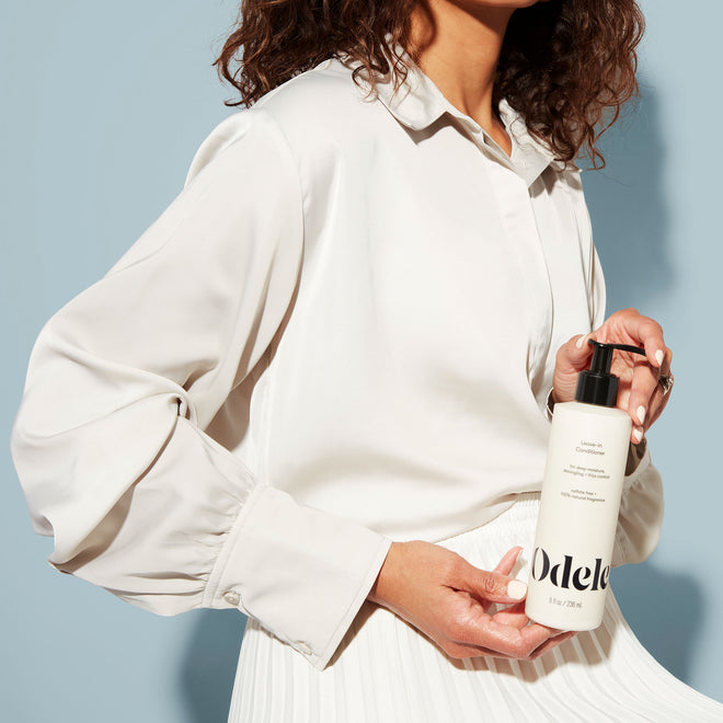 A woman in a white blouse and skirt holds a bottle of Odele Leave-in Conditioner with both hands against a plain, light blue background. Her face is not shown.