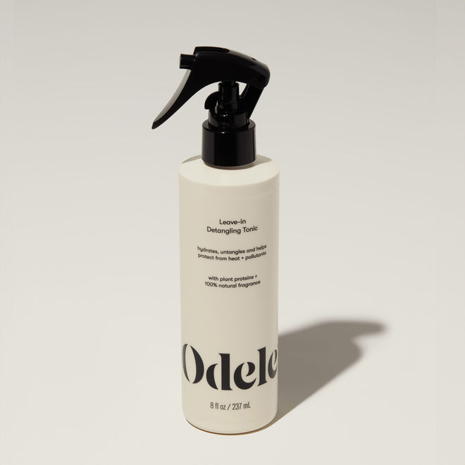 A bottle of Odele Leave-in Detangling Tonic stands upright against a plain, light-colored backdrop with the front of the label facing the camera.