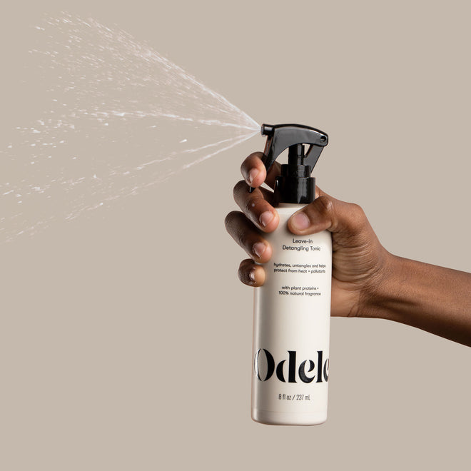 A brown-skinned hand sprays Odele Leave-in Detangling Tonic in front of a plain beige background.