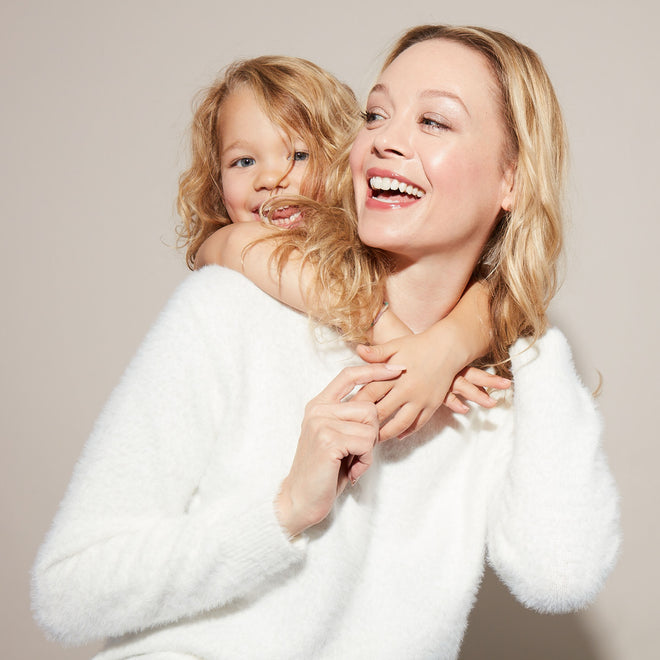 A smiling woman in a white sweater carries a laughing blonde girl on her back, both happy against a neutral background.
