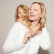 A smiling woman in a white sweater carries a laughing blonde girl on her back, both happy against a neutral background.