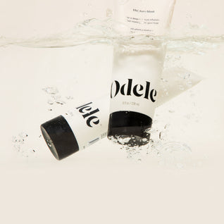 Two white tubes of Odele Moisture Mask Mini enriched with argan and jojoba are partially submerged in clear water, creating bubbles and ripples around this nourishing, hydrating hair treatment.