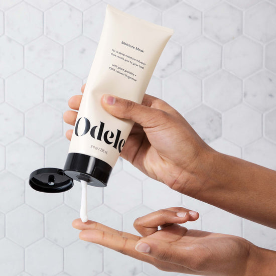 Someone dispenses white cream from a tube of Odele Moisture Mask onto their finger in front of a grey hexagonal tiled wall in the background.
