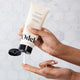 Someone dispenses white cream from a tube of Odele Moisture Mask onto their finger in front of a grey hexagonal tiled wall in the background.