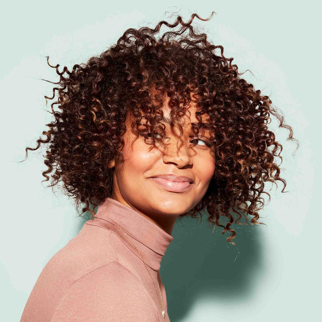 A woman with medium-brown skin and short, curly hair smiles softly while looking over her shoulder in front of a plain light green background.