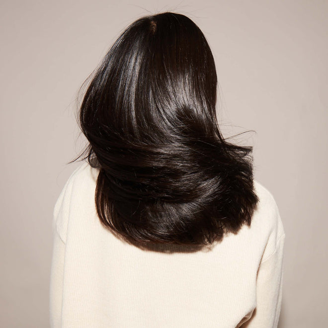 A person with shoulder-length, shiny black hair faces away from the camera and swishes their hair back and forth in front of a plain beige background.
