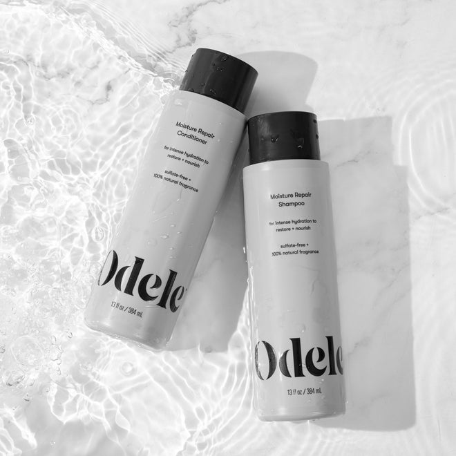A bottle of Odele Moisture Repair Shampoo and Conditioner rest on a wet surface with water ripples.