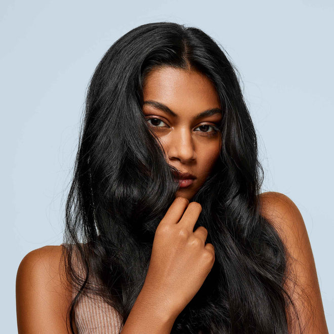 A woman with long, wavy black hair poses against a light blue background.