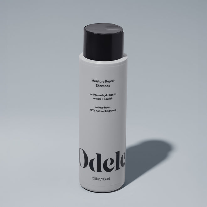 A bottle of Odele Moisture Repair Shampoo stands upright against a plain, light-colored backdrop with the front of the label facing the camera.