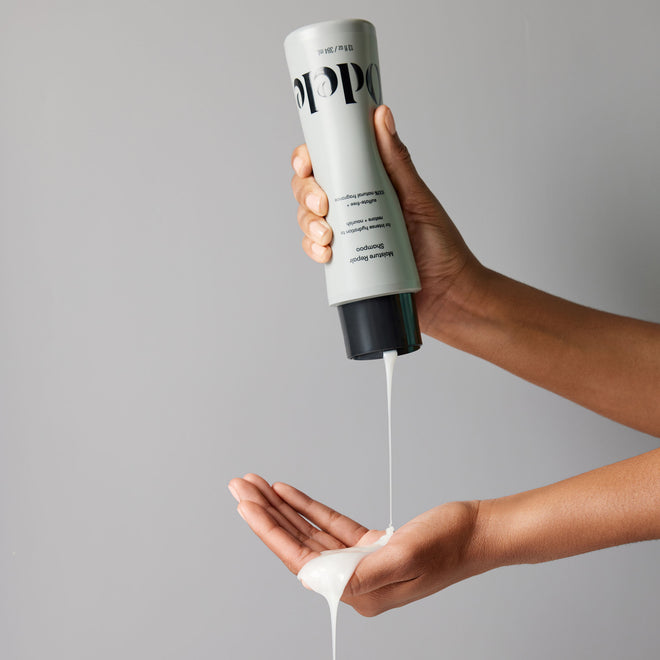 A person squeezes Odele Moisture Repair Shampoo from a gray bottle into their palm, set against a plain gray background.