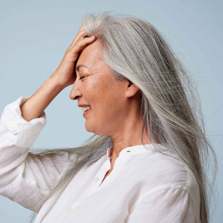 A smiling older woman with long gray hair in a white shirt touches her forehead, radiating confidence against a light blue background—she looks like she’s just discovered Odele Beauty’s Moisture Repair Shampoo Mini for restoring brittle hair.