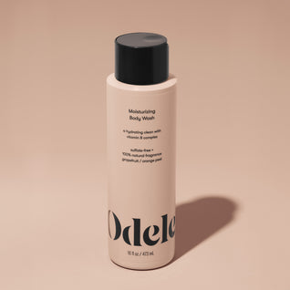A bottle of Odele Moisturizing Body Wash stands upright against a plain, light orange backdrop with the front of the label facing the camera.