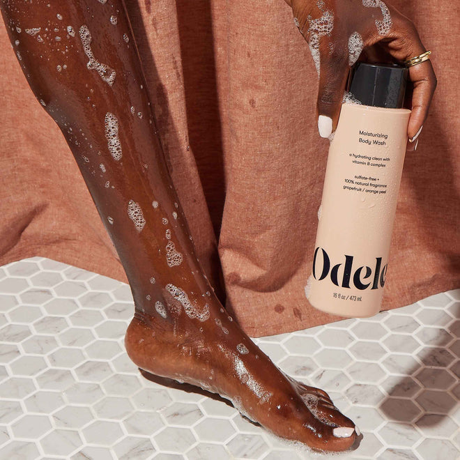 A person with wet, soapy skin stands on white hexagonal tiles, holding a bottle of Odele Beauty Moisturizing Body Wash for a hydrating clean. A brown fabric backdrop hangs behind them.