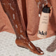 A person with wet, soapy skin stands on white hexagonal tiles, holding a bottle of Odele Beauty Moisturizing Body Wash for a hydrating clean. A brown fabric backdrop hangs behind them.
