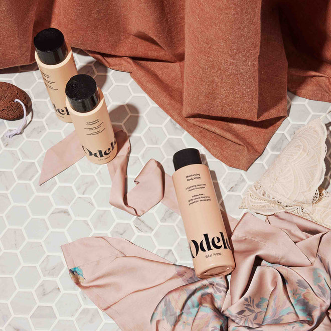 Three bottles of Odele Moisturizing Body Wash rest on a white hexagonal tile floor next to a pink floral scarf, pink bra, and rust-colored fabric.
