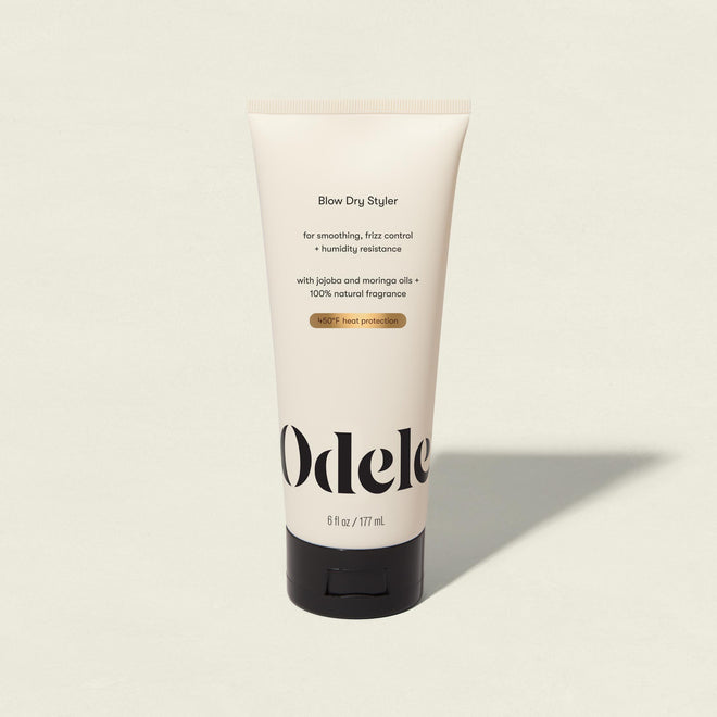 An upright beige tube of Blow Dry Styler rests on a light surface, its label with black and gold text emphasizing frizz control, humidity resistance, and heat protection against a minimalist background.