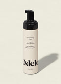 A beige bottle of Curl Defining Foam with a black pump top, labeled for lightweight, bouncy hold and frizz control, stands on a neutral background.