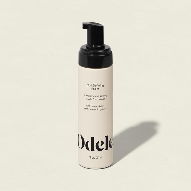 A beige bottle of Curl Defining Foam with a black pump top, labeled for lightweight, bouncy hold and frizz control, stands on a neutral background.