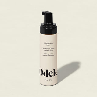 A beige bottle of Curl Defining Foam with a black pump top, labeled for lightweight, bouncy hold and frizz control, stands on a neutral background.