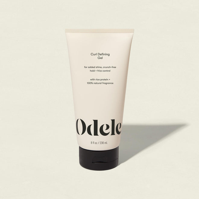 An upright tube of Curl Defining Gel stands on a light background. Its cream-colored packaging displays black text, highlighting product benefits and ingredients like rice protein and natural fragrance.