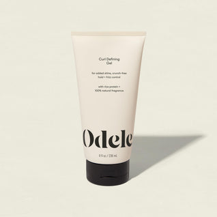 An upright tube of Curl Defining Gel stands on a light background. Its cream-colored packaging displays black text, highlighting product benefits and ingredients like rice protein and natural fragrance.